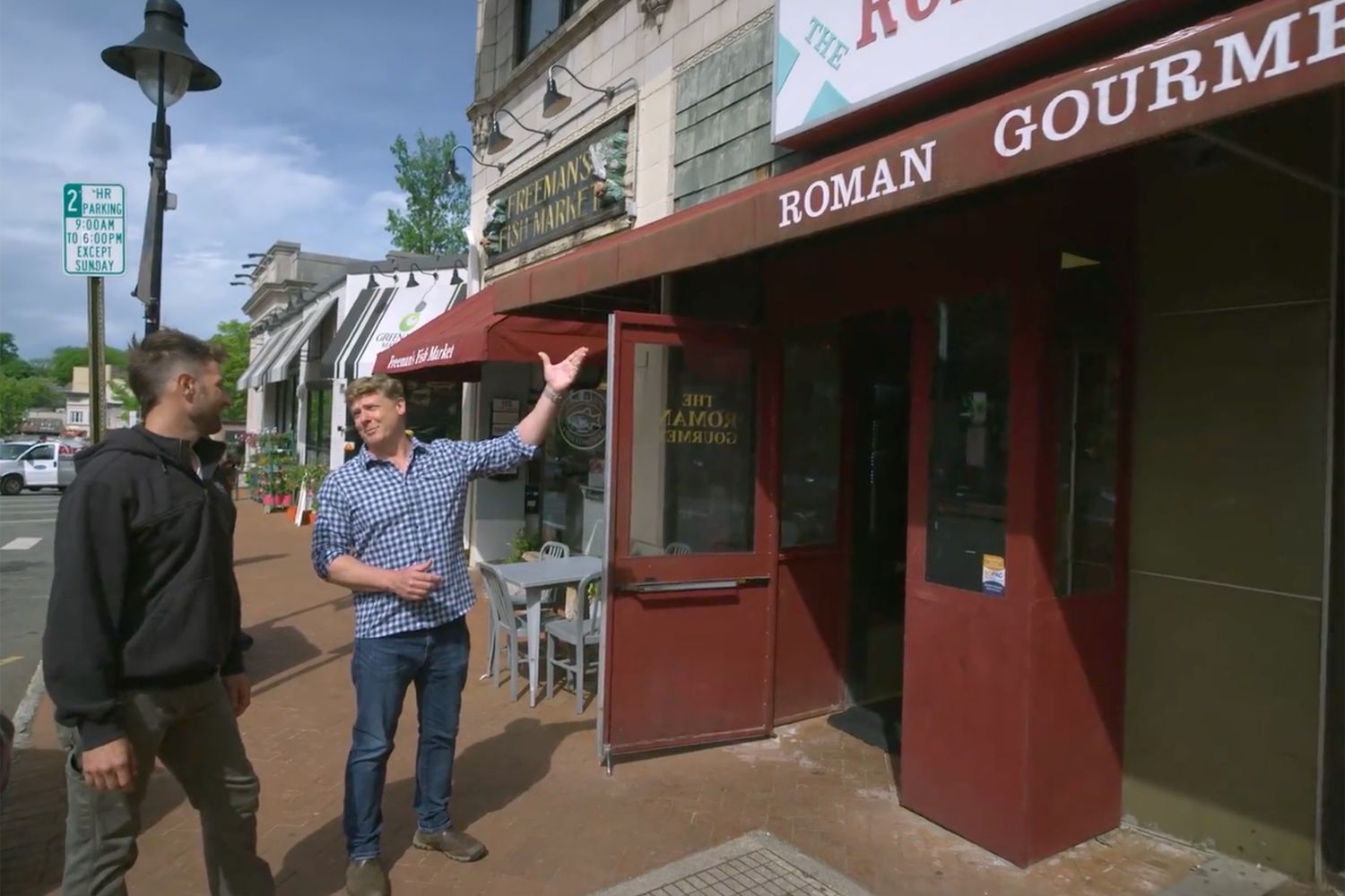 Two people stand outside Roman Gourmet, a red storefront on a street with other shops and outdoor seating. One person gestures upward. A streetlamp and parking sign are visible in the background.